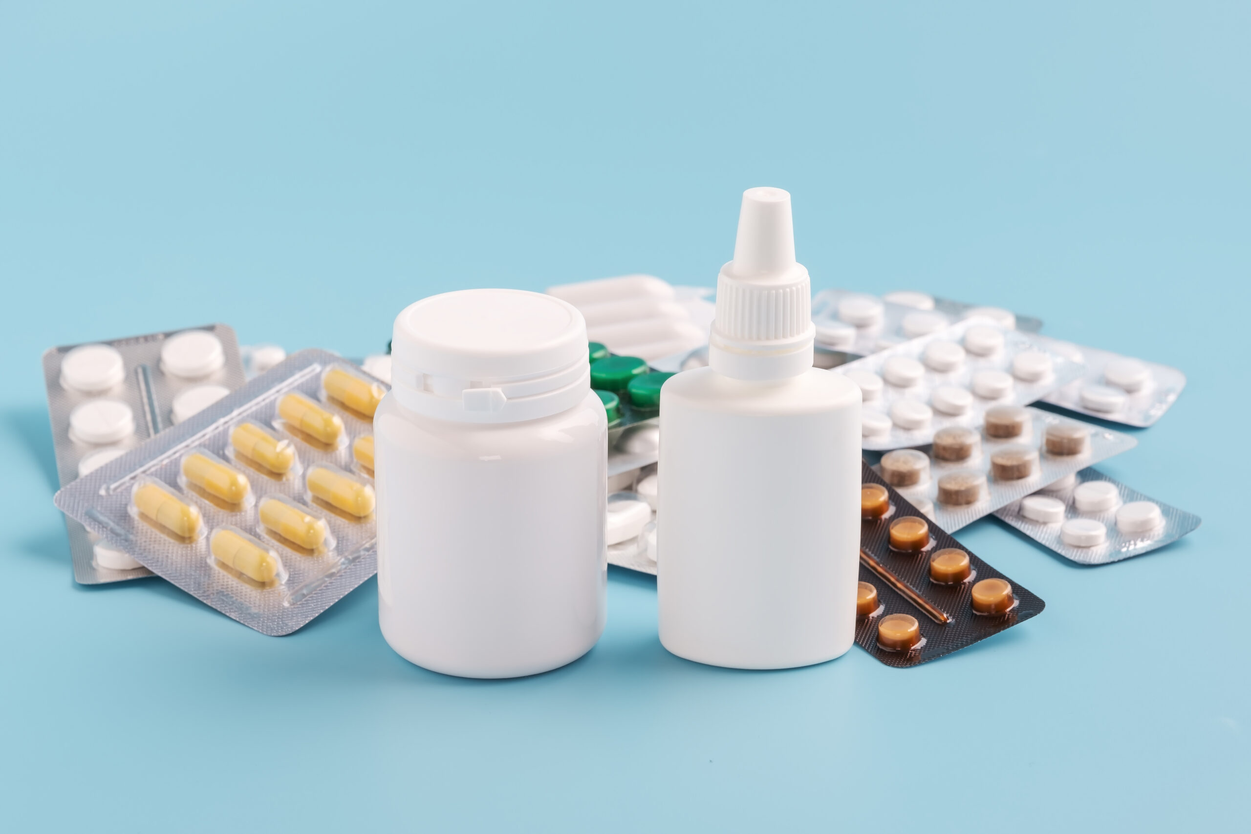 Two white bottles foreground and pile of various shapes and sizes of drugs in different packaging on a blue background.