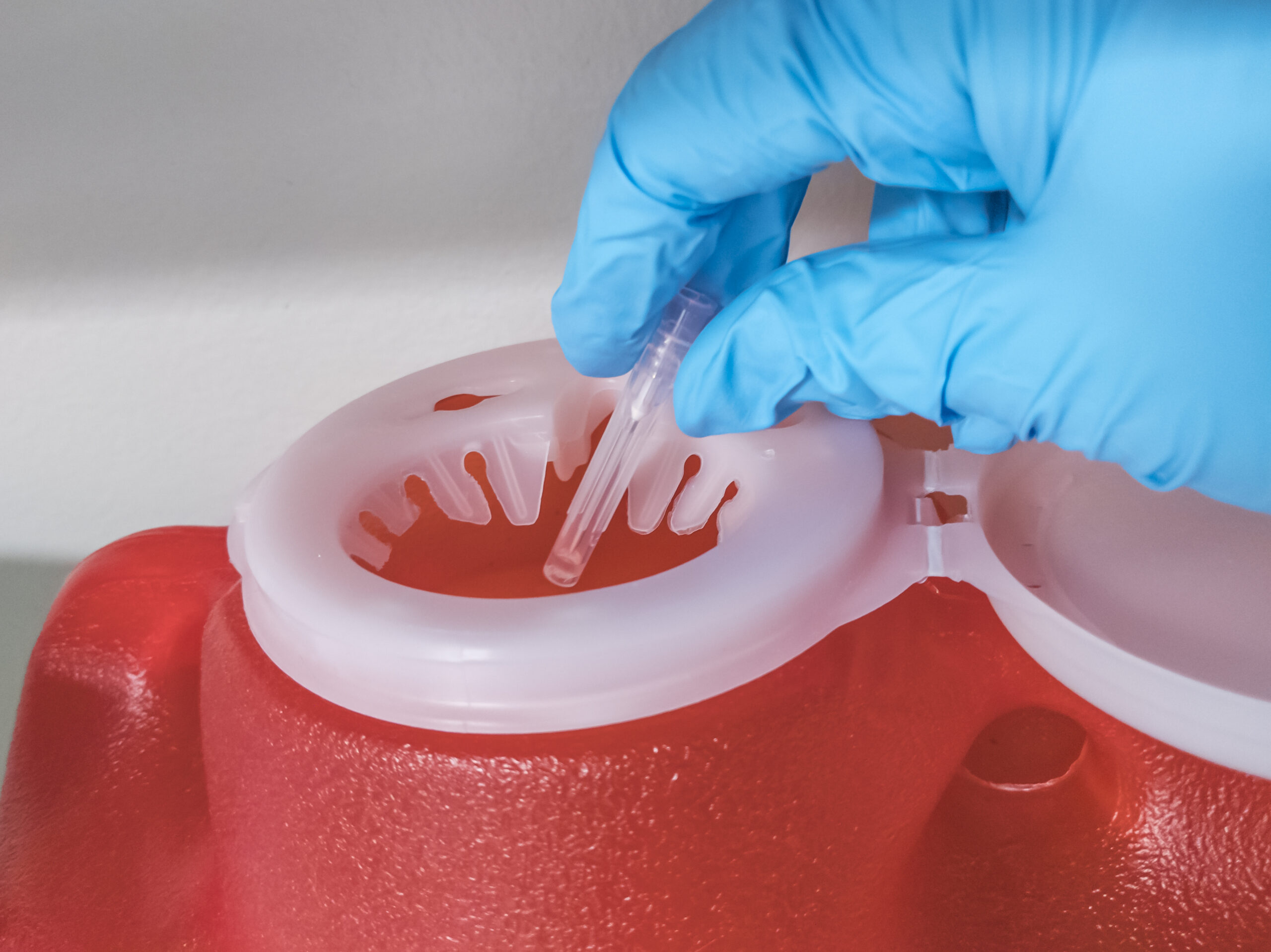 Close up of a sterile, gloved hand dropping a used needle into a bright red sharps container in a medical facility